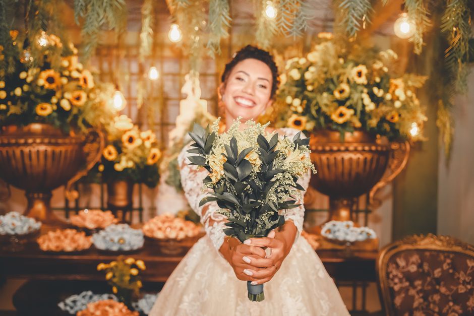 A smiling woman in a white lace wedding dress holds a bouquet of fresh flowers featuring dark green foliage and delicate cream-colored blooms. She stands in a warmly lit room decorated with lush flora