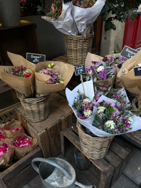 An outdoor display of fresh flower bouquets arranged in woven wicker baskets, each wrapped in light pink or brown paper with small chalkboard signs indicating prices, such as 29€ and 26€. The flow