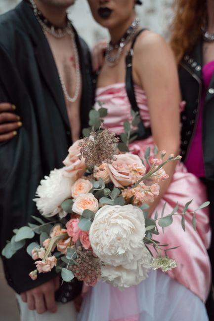 A close-up of a floral bouquet featuring large, soft-white peonies, blush pink roses, and delicate pink ranunculus, accented with eucalyptus leaves and small filler flowers. The arrangement is held by