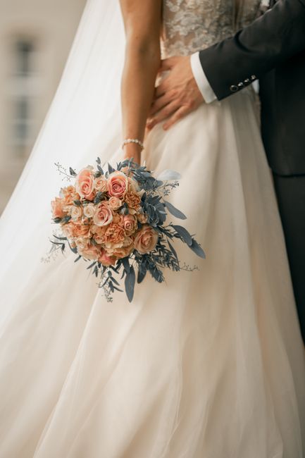 A bride holding a floral bouquet consisting of blush pink roses, peach-colored spray roses, and sprigs of dusty blue eucalyptus leaves, with the stems wrapped in a soft ribbon. The bouquet is position